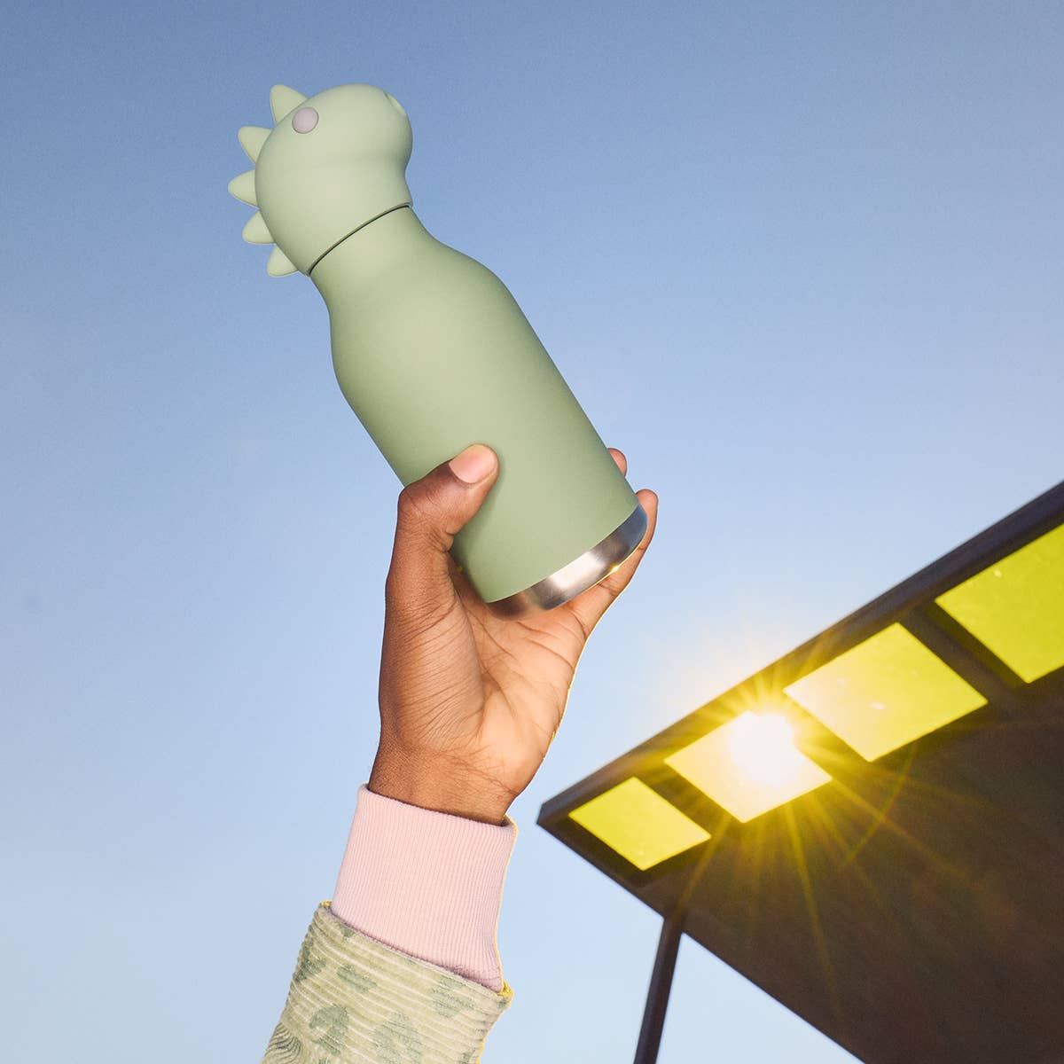 Person holding a green dinosaur-shaped water bottle with a solar panel in the background