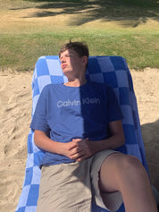 Person sitting on a blue checkered blanket outdoors on the sand with the shadow of palm trees on the grass nearby