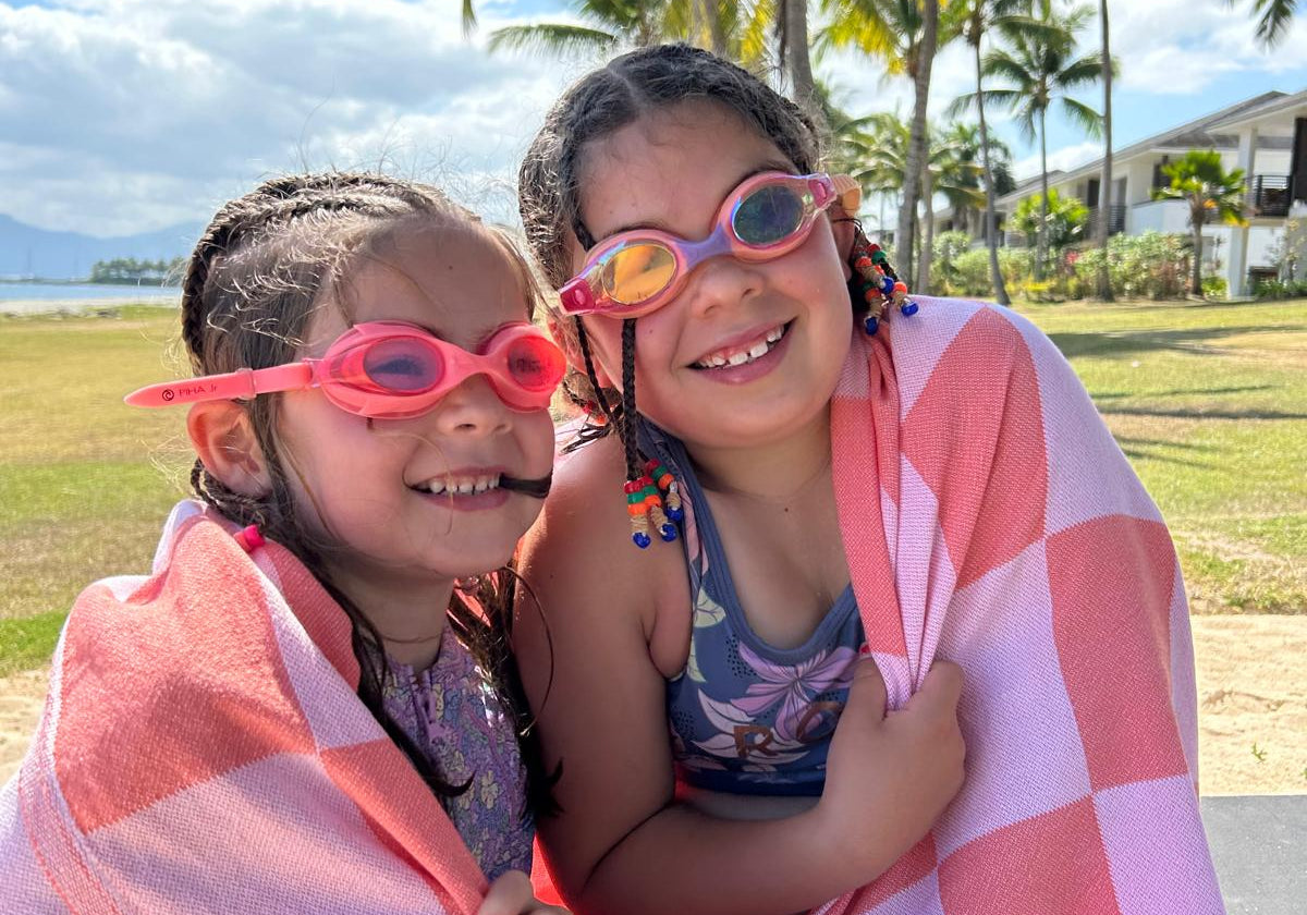Two children wrapped in a pink towel on a beach with palm trees in the background