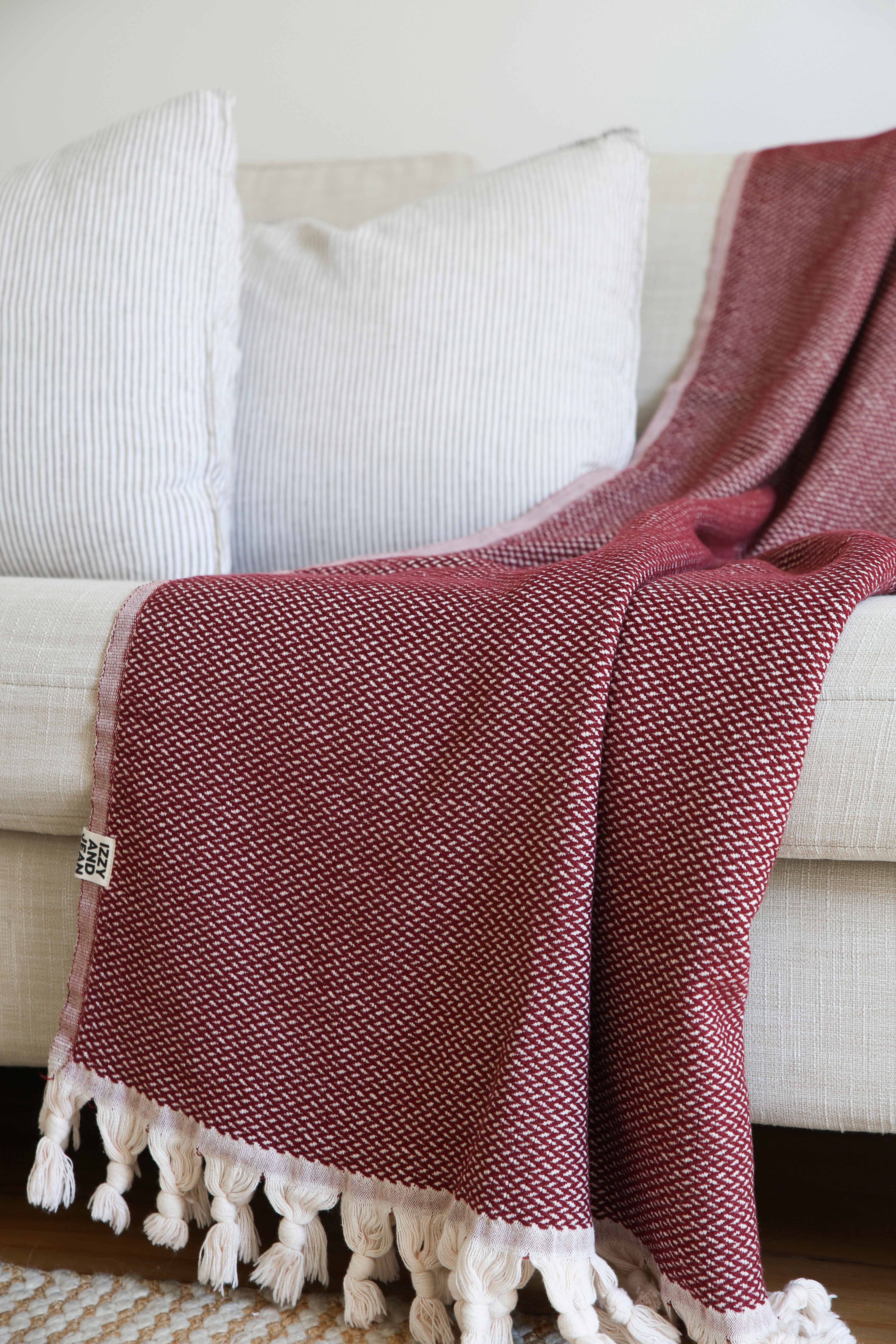A burgundy throw blanket made from 100% cotton, with a herringbone pattern and fringed edges on a white background on a white couch in a living room.