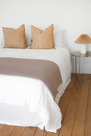 Bed with beige pillows, white bedspread with the brown riveria throw blanket across the bed