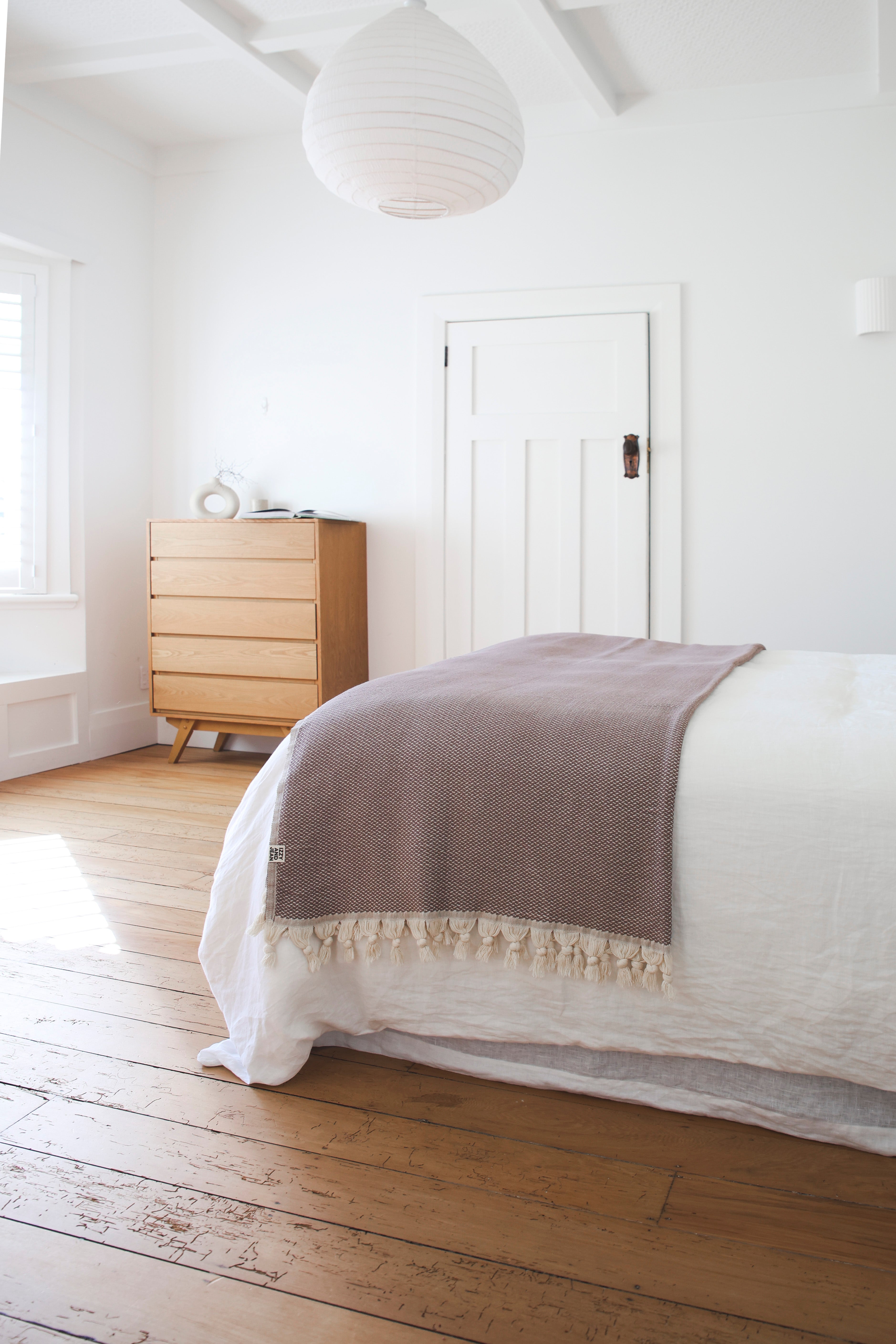 Bed with a brown riveria throw blanket in a room with wooden flooring and a dresser.