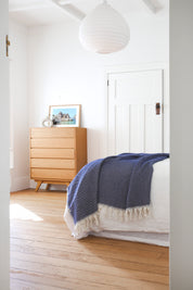Bedroom with a wooden dresser and The Riveria Dark Blue throw blanket with tassels on the bed.