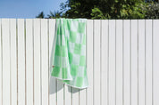 Green and white checkered towel hanging on a white wooden fence with trees in the background.