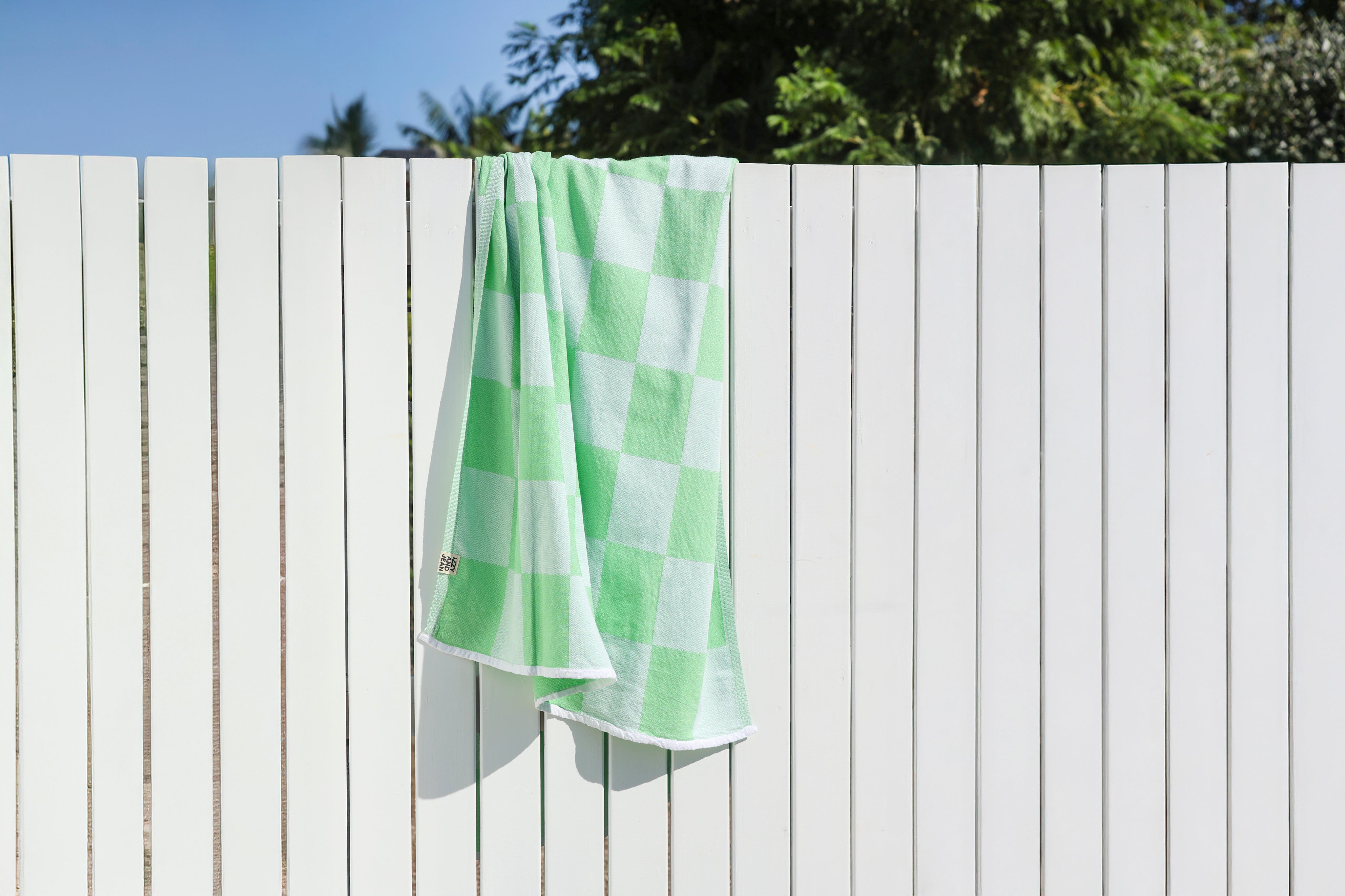 Green and white checkered towel hanging on a white wooden fence with trees in the background.