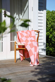 A coral and white checkered towel draped over a wooden chair on a wooden deck with a white building and greenery in the background.