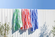 Three colourful Checkered Turkish Towels in coral, royal blue and apple green towels hanging on a clothesline against a white fence with a clear blue sky