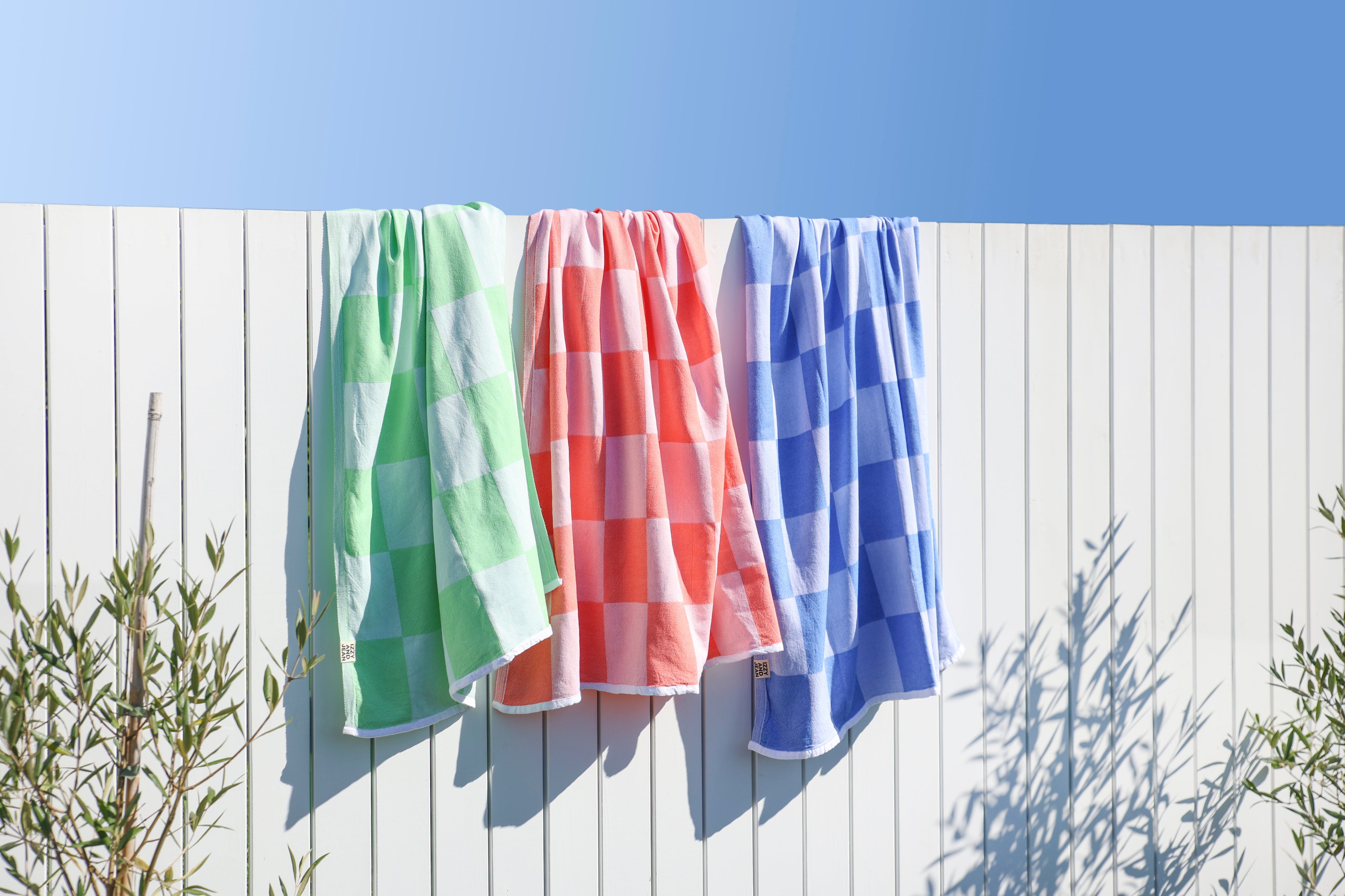 Three colourful Checkered Turkish Towels in coral, royal blue and apple green towels hanging on a clothesline against a white fence with a clear blue sky
