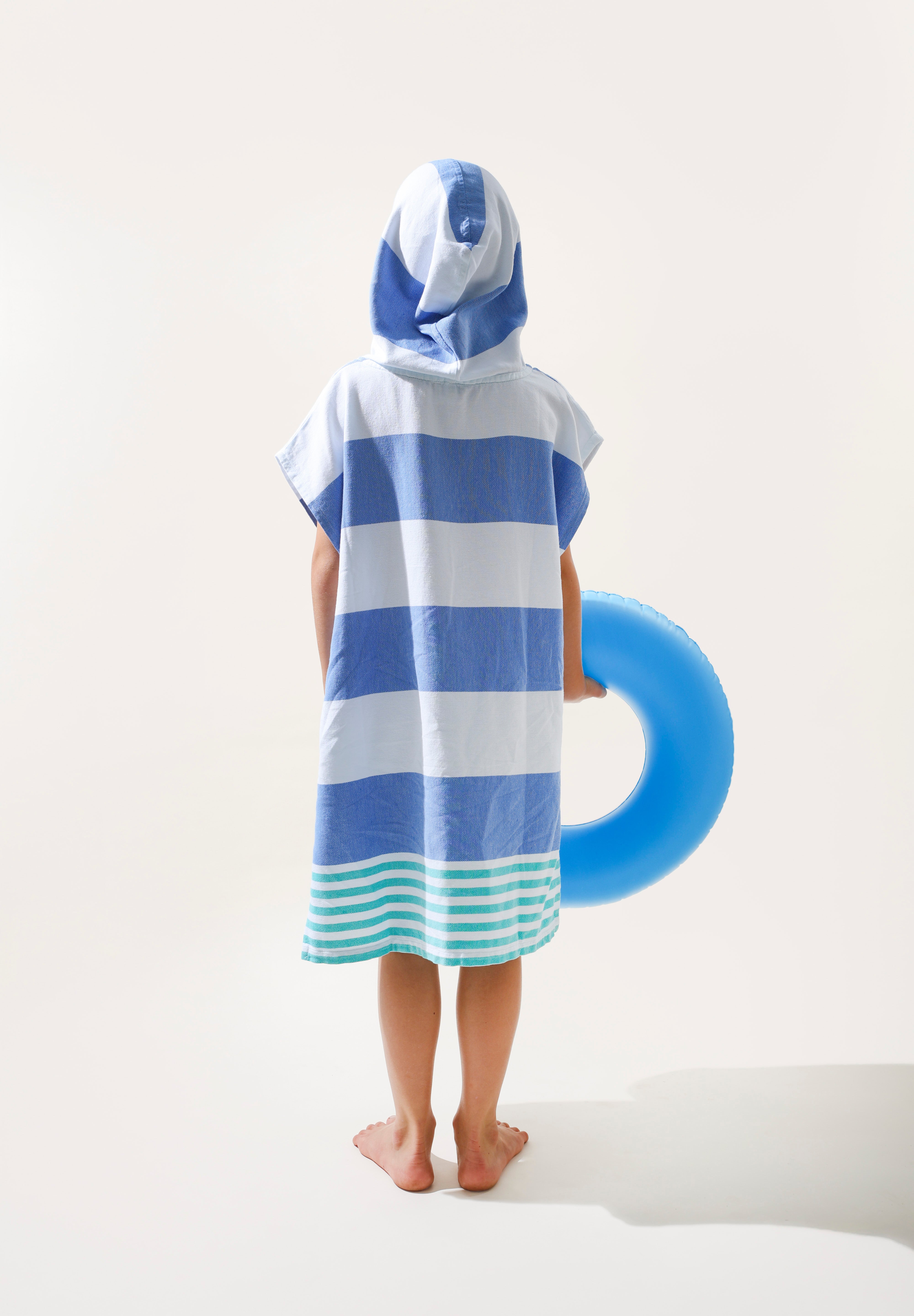 Child wearing a blue and green striped ezy-towel with a hood, made from cotton. Standing next to a blue inflatable ring on a white background.