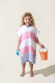 Child wearing a pink and white striped hooded Turkish towel, holding a blue toy and an orange bucket on a beige background