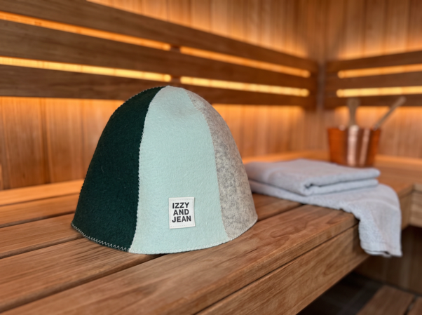 A dome-shaped knit cap with three color-blocked panels in dark green, pale mint, and biscotti, featuring a white square label that reads 'IZZY AND JEAN'. It is displayed on a wooden sauna bench next to a folded light blue towel.