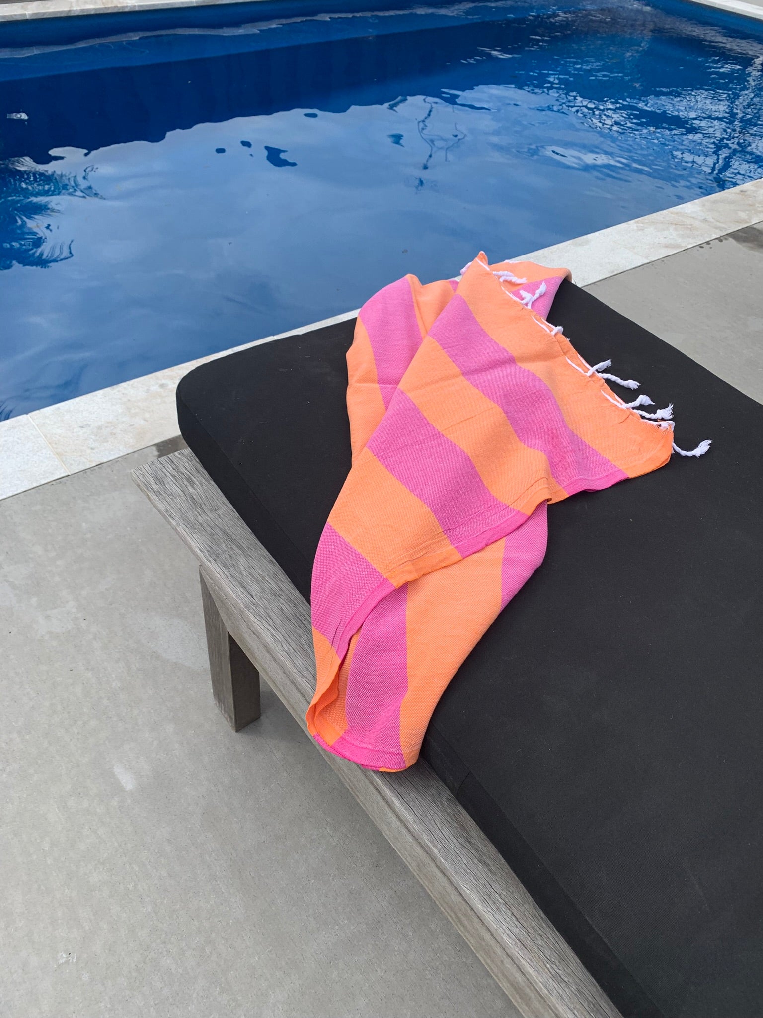 Orange and hot pink Turkish Towel with white tassels on sun bed by a pool.