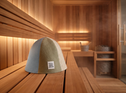 A dome-shaped felt sauna hat by Izzy and Jean wool hat with three color-blocked panels in ice blue, pale mint, and biscotti sits on a wooden sauna bench inside a wooden sauna room with warm ambient lighting.