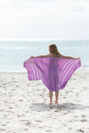 a person on the beach facing the ocean with the purple Turkish towel outstretched in her arms behind her