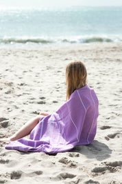 A little girl wrapped in the Classic Purple Turkish towel sitting on the sand looking at the waves