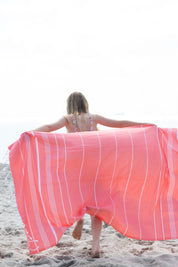A girl on a beach running away from the camera with her arms outstretched and the Classic Turkish towel in coral with white stripes flowing behind her.