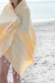 A yellow Turkish towel with white stripes and fringes on the bottom wrapped around a girl on the beach