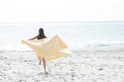 A yellow Turkish towel with white stripes and fringes on the bottom bellowing out behind a girl running away from the camera towards the ocean on a sand