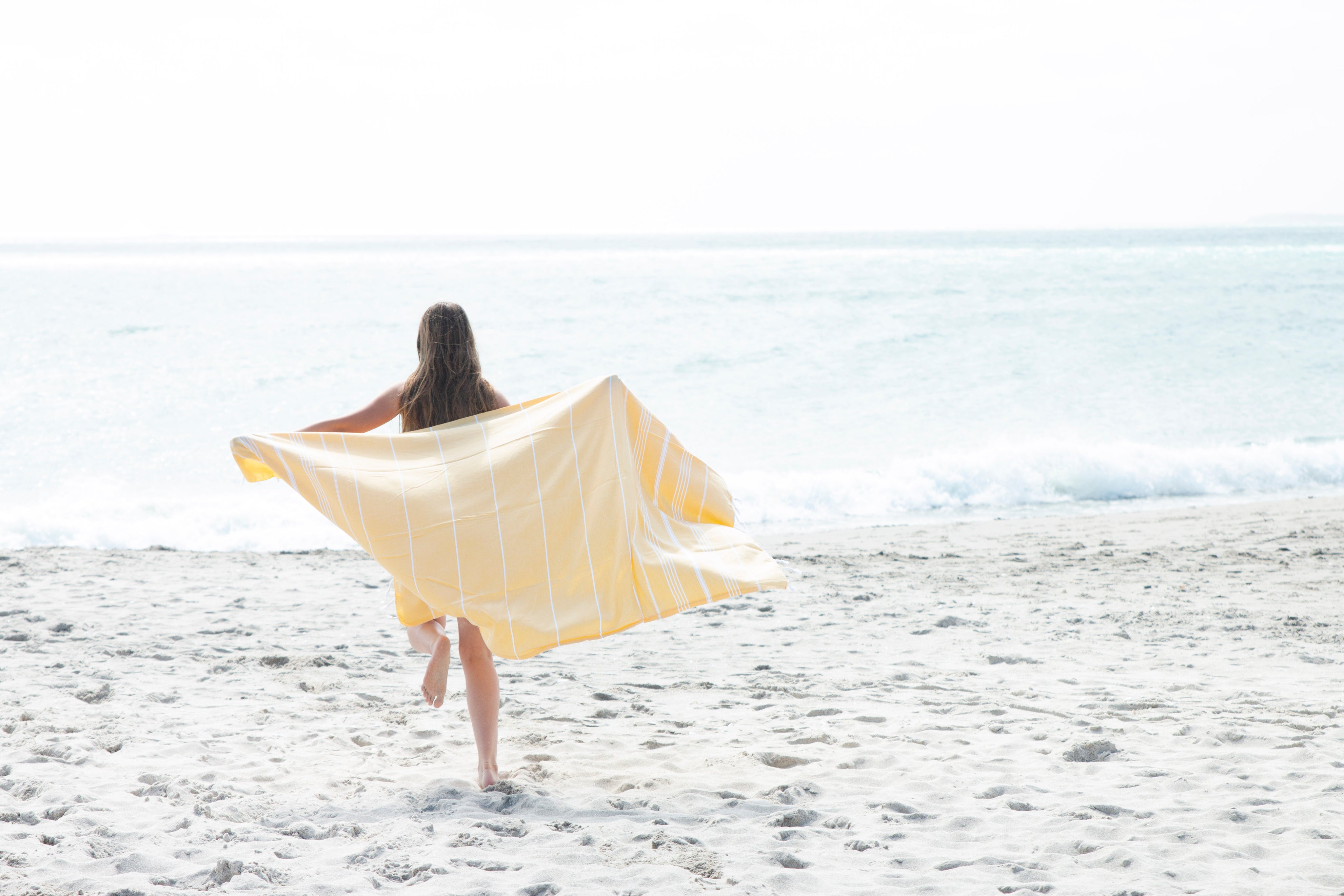 A yellow Turkish towel with white stripes and fringes on the bottom bellowing out behind a girl running away from the camera towards the ocean on a sand