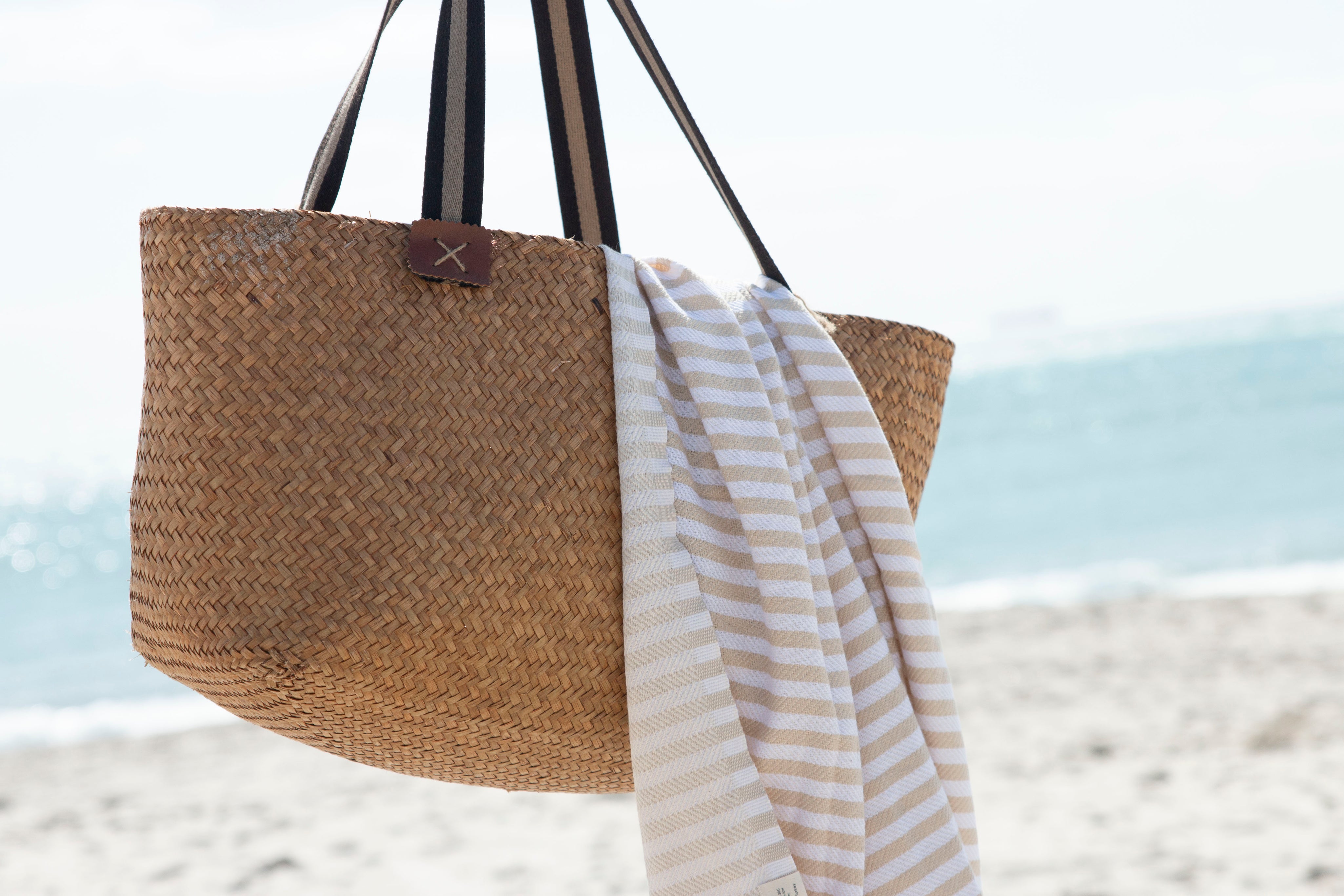 A St Tropez sand and white stripped 100% cotton towel hanging out of a beach bag with the beach in the background