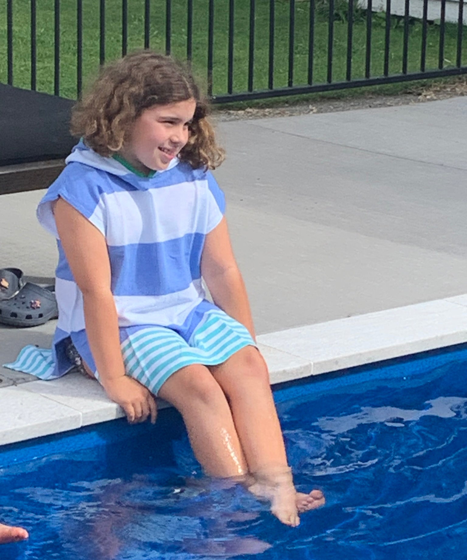 Child sitting on side of a pool with legs in water, wearing the Ezy-towel hooded 100% cotton towelie in blue and green. 
