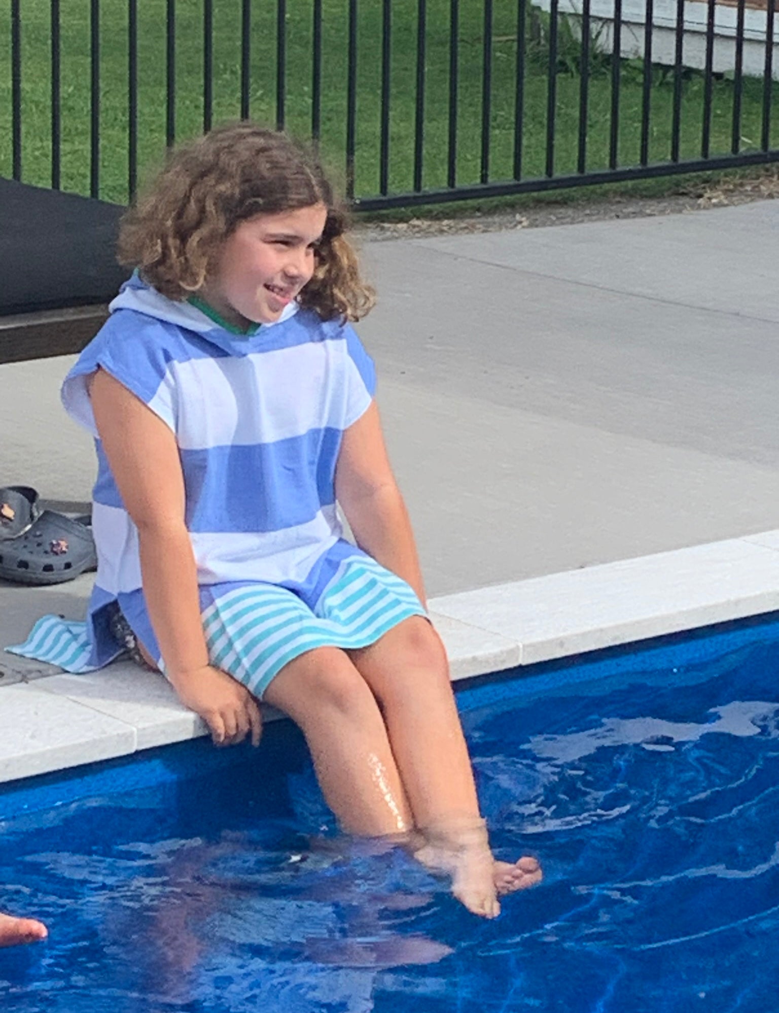 Child sitting on side of a pool with legs in water, wearing the Ezy-towel hooded 100% cotton towelie in blue and green. 