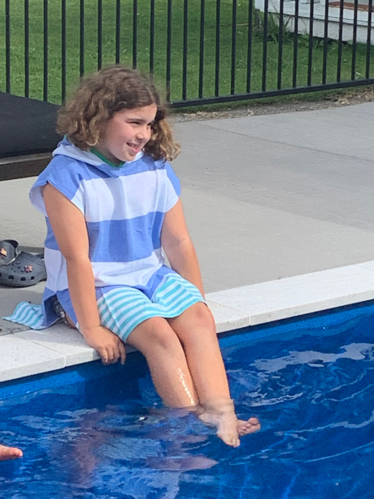 Child sitting on side of a pool with legs in water, wearing the Ezy-towel hooded 100% cotton towelie in blue and green. 