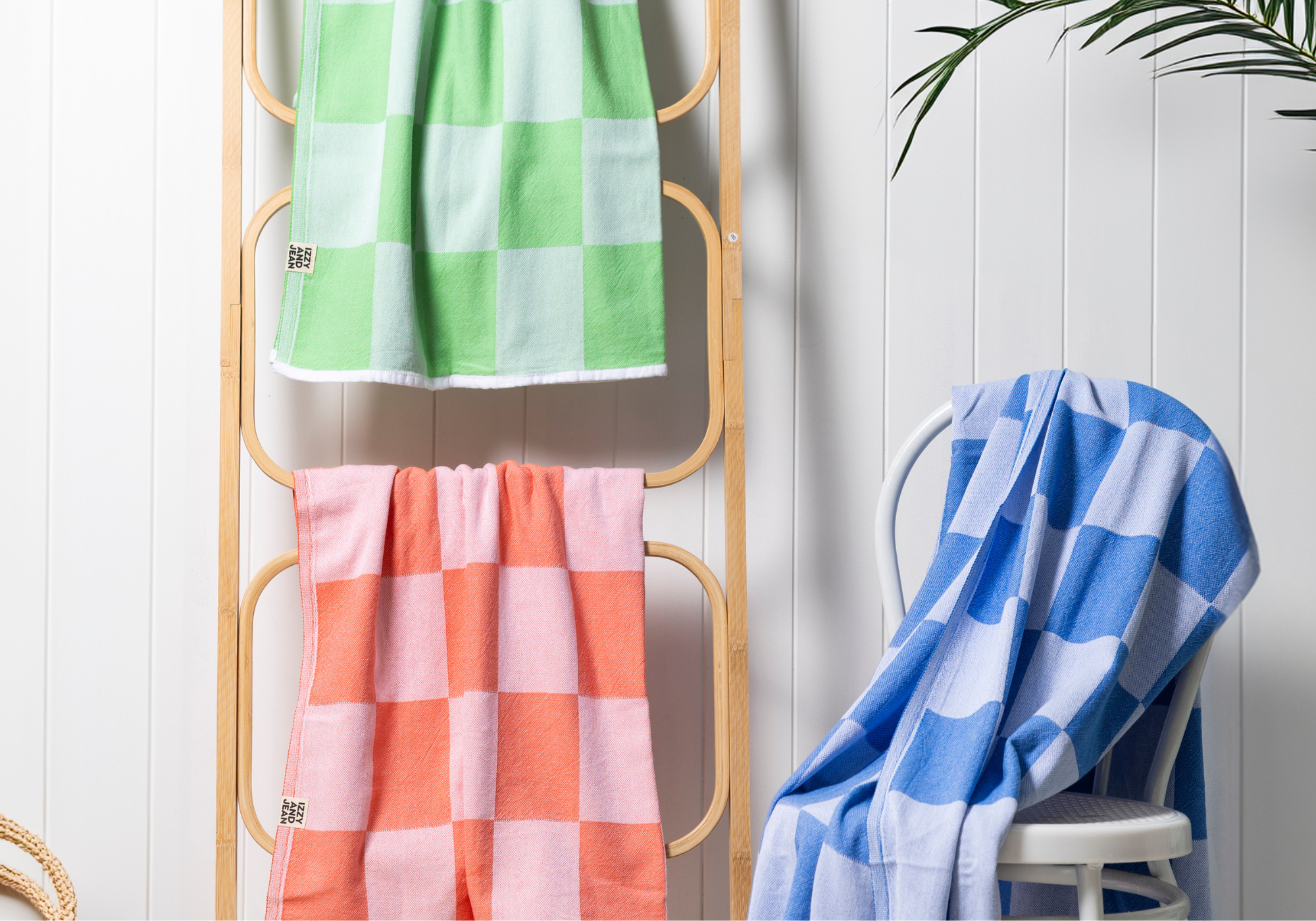 apple green, coral and blue checkered Turkish towels hanging on a ladder shelf and white wooden chair with a wooden white wall behind them and plant in the corner.