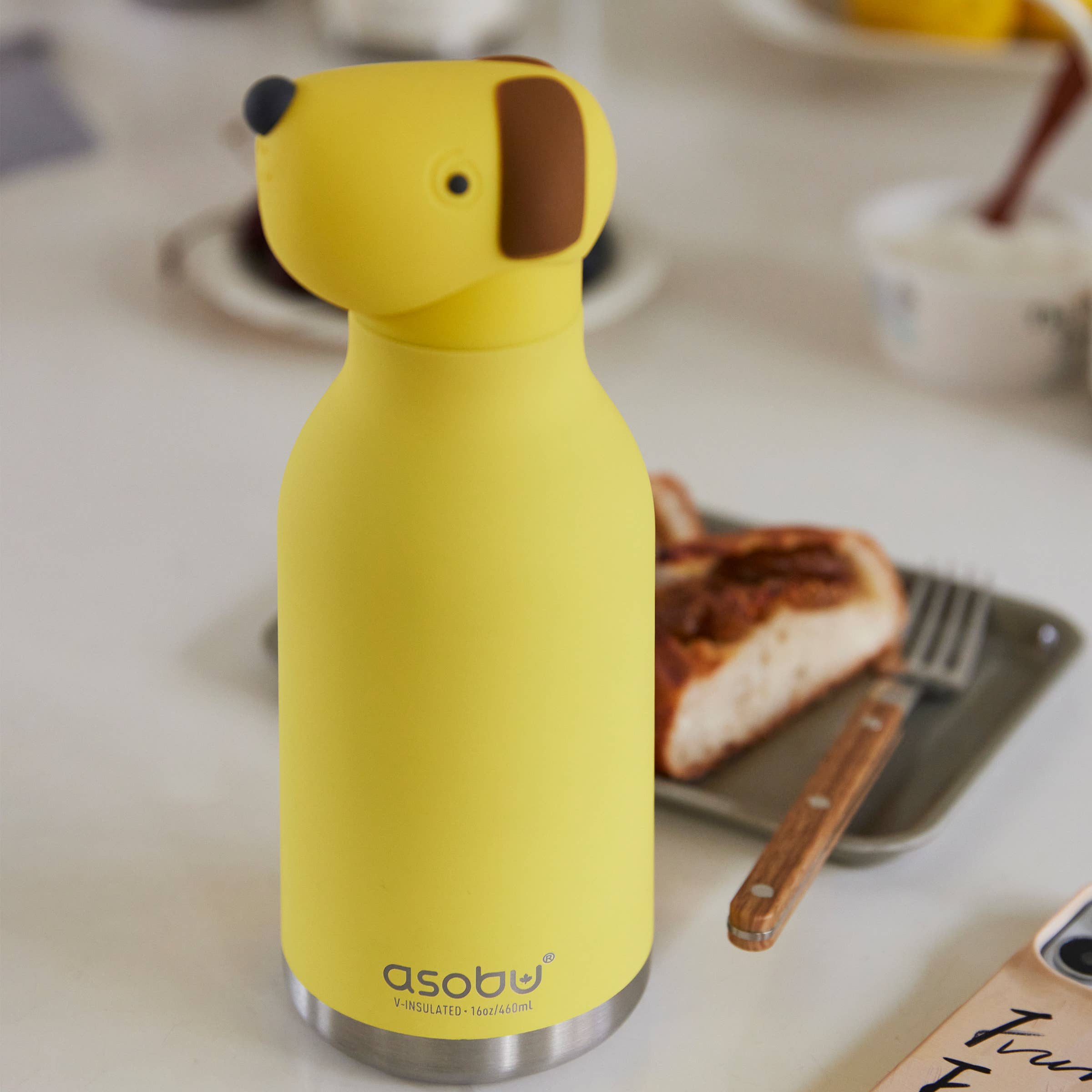 Yellow dog-shaped water bottle on a table with toast and a plate in the background
