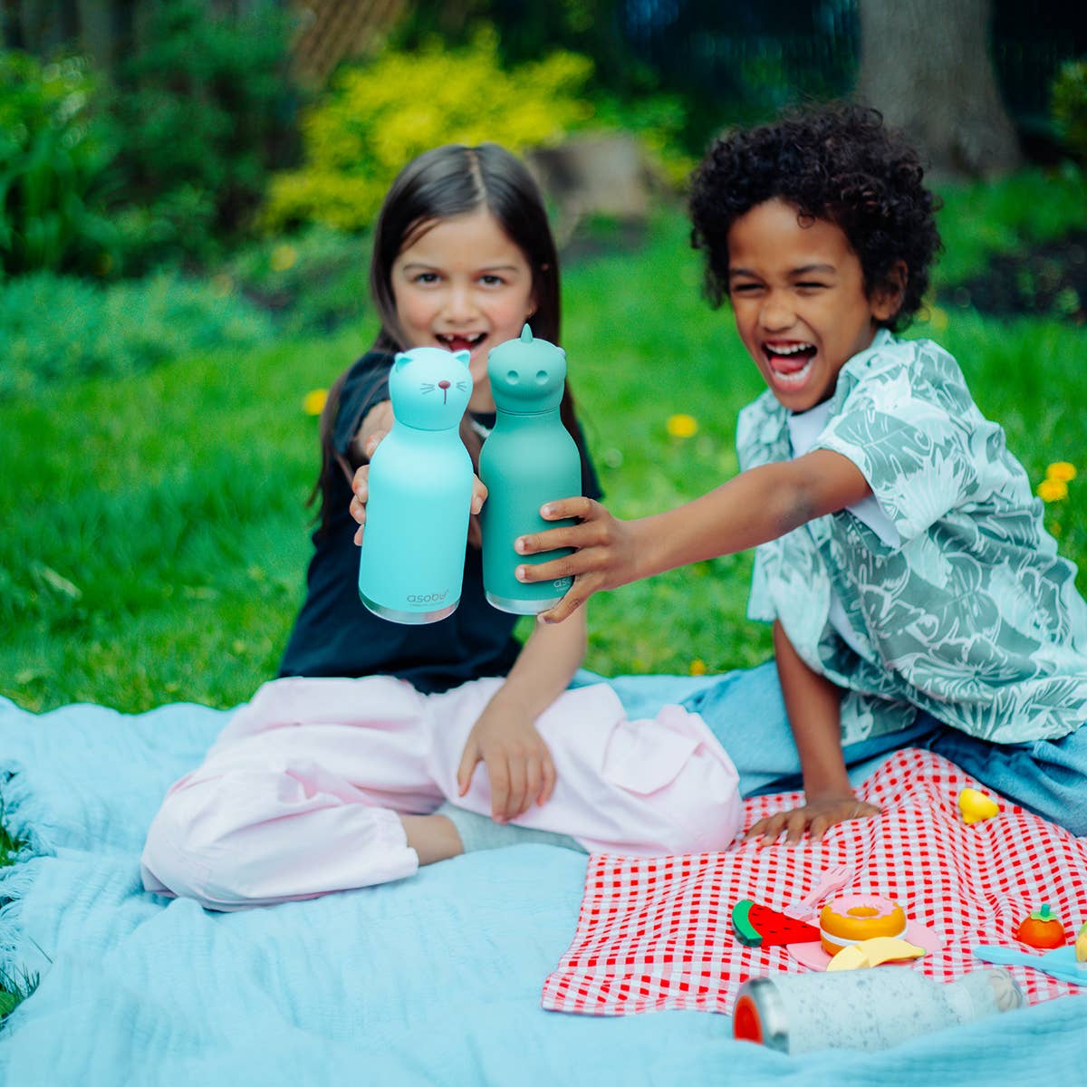 Two children playing with blue cat-shaped bottles on a picnic blanket outdoors.