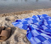 A checkerboard-patterned towel in royal blue, laid out on sand beside a pair of brown sandals, with a body of water in the background.