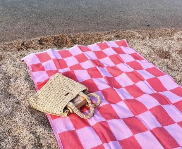 A coral and white checkerboard-patterned Turkish towel laid out on the sand with a beige tote bag on top of it, near a body of water.