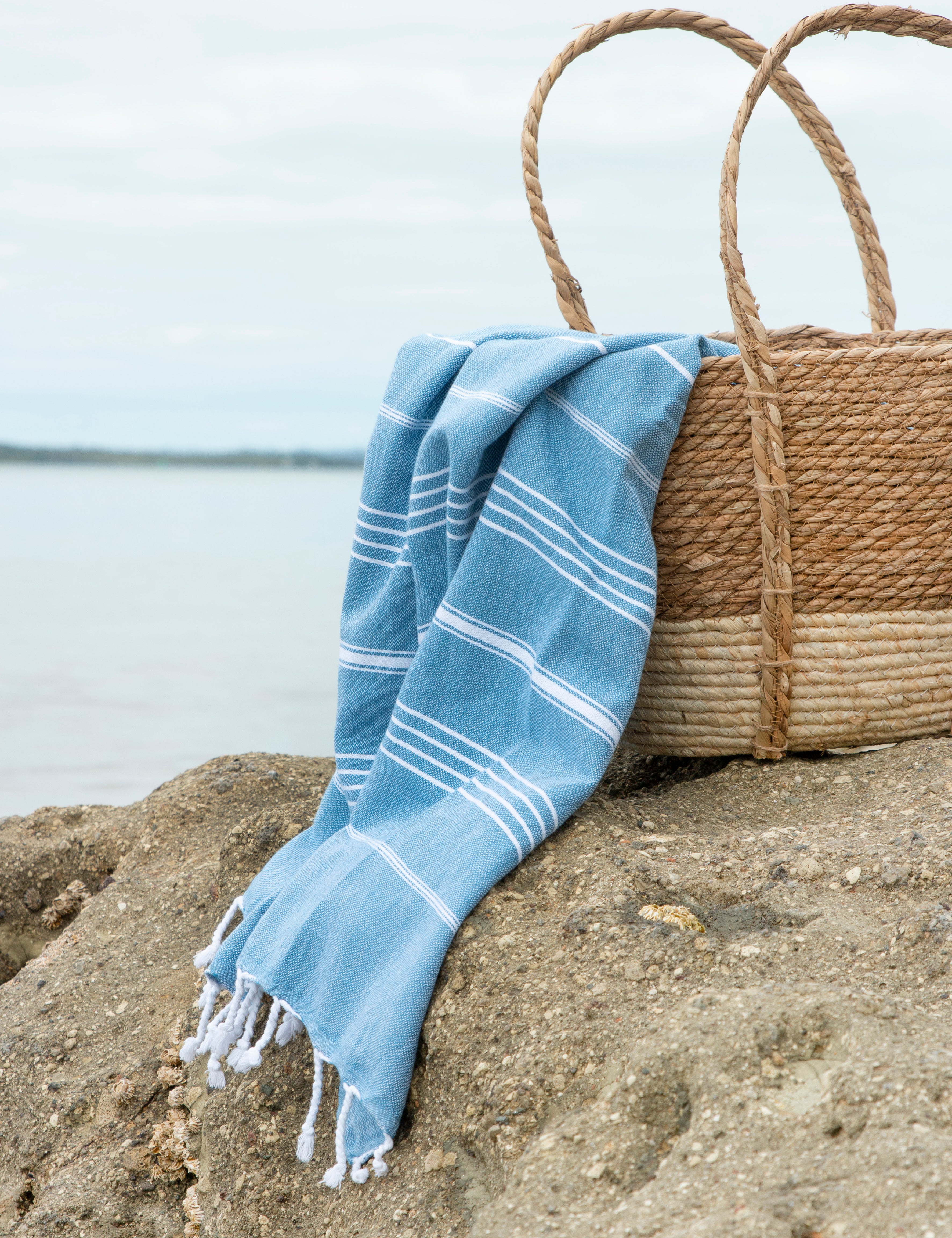 The Aegean Blue Turkish Towel in a weave basket on the sand with the sea in the background