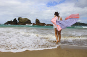 a lady in a white dress playing in the shallow waves on the beach with a blue and coral striped Turkish towel bellowing out behind her.