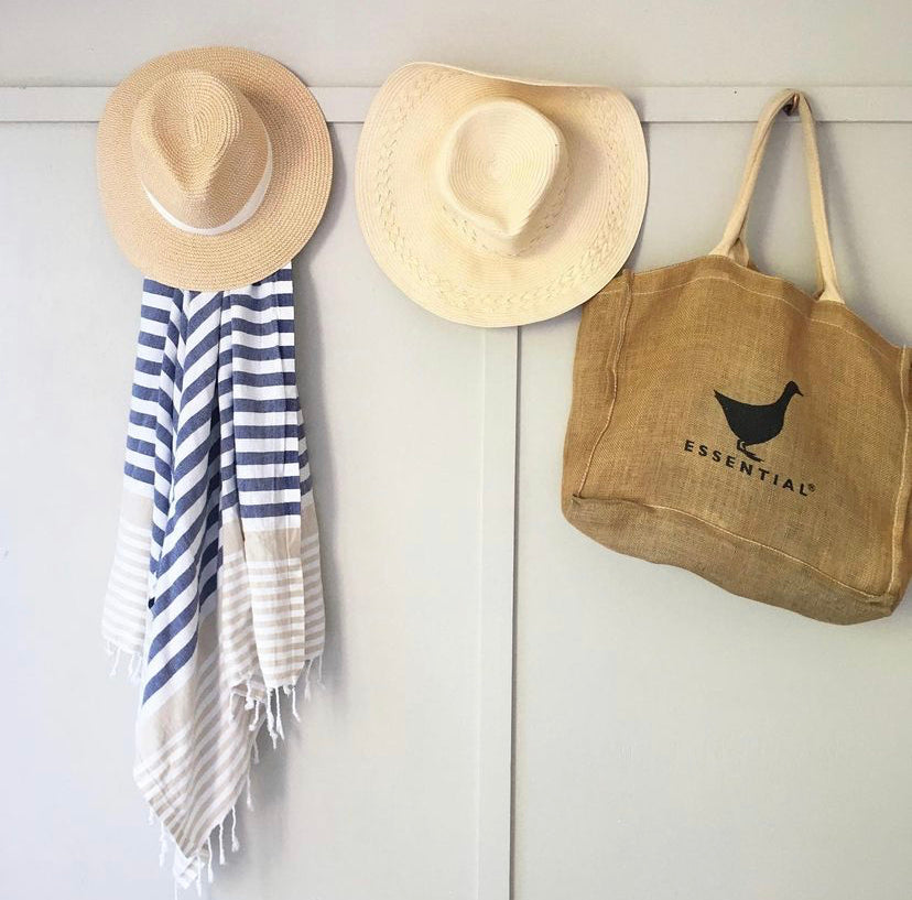 A Navy and Sand striped Sofia Turkish Towel with tassles at the bottom hanging from a hook on a wall, with 2 sun hats and a beach bag on the other hooks next to it