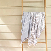 A pale grey Turkish towel with white stripes and tassels on the bottom edge hangs on a wooden ladder against a mustard coloured wall