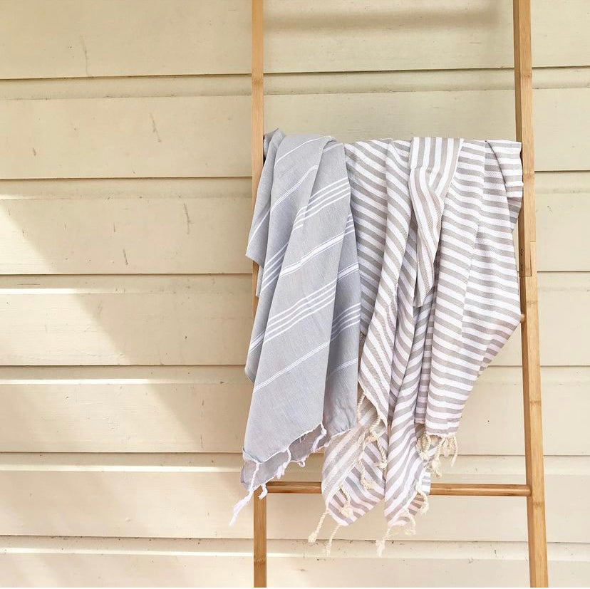 A pale grey Turkish towel with white stripes and tassels on the bottom edge hangs on a wooden ladder against a mustard coloured wall