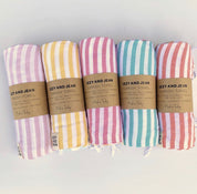 5 rolled Turkish Towels in a row with an Izzy and Jean branded paper wrap around each one, each towel is a different colour and white stripe starting with purple, yellow, fuschia, ocean and coral.
