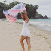 a girl playing in the wind on the beach wearing a white dress holding up a blue and coral stripped Turkish Towel flowing in the wind above her.