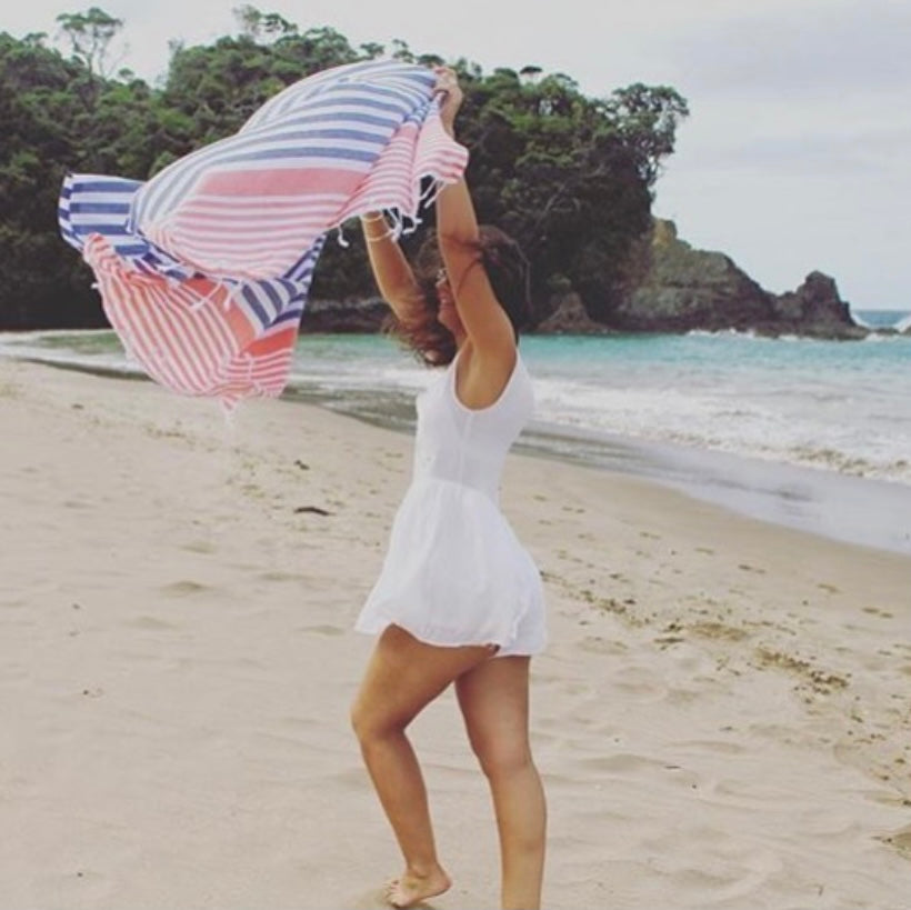 a girl playing in the wind on the beach wearing a white dress holding up a blue and coral stripped Turkish Towel flowing in the wind above her.