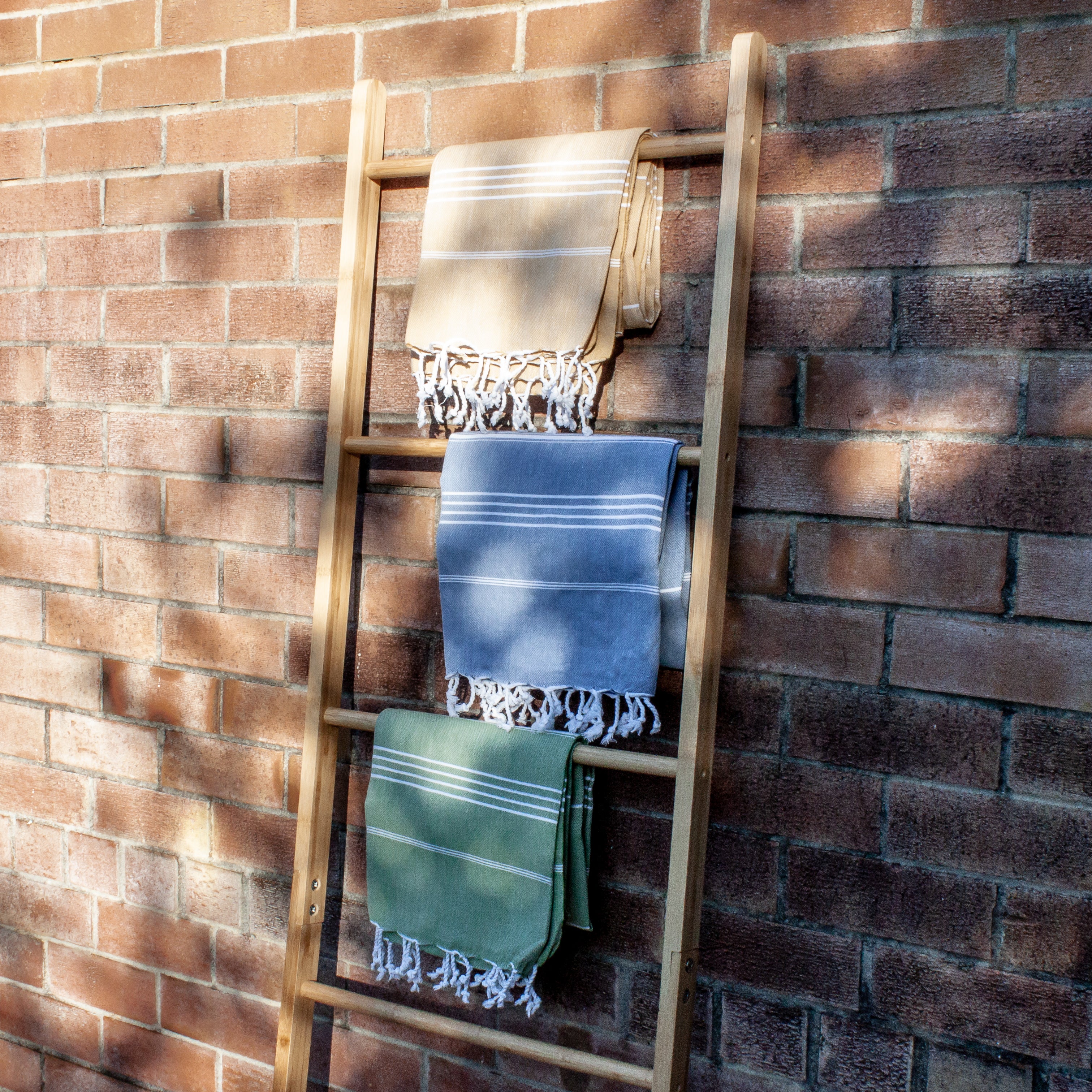 Three Turkish towels with white stripes and fringes on the bottom, folded over a wooden ladder with a brick background in olive and yellow and navy