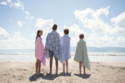 4 children standing on a beach, facing the water except the one and only girl on the left smiling at the camera wrapped in a coral and fuchsia striped Turkish beach towel 