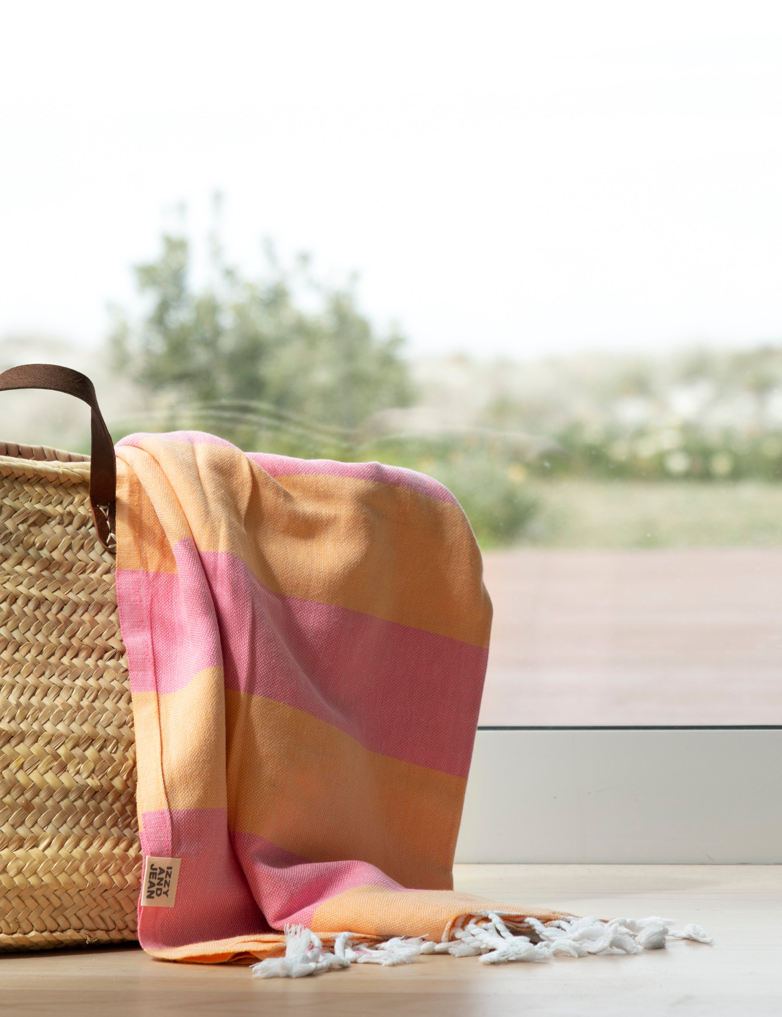 The striped Santorini Turkish towel beach towel in melon and pink hangs over a weave basket near a window overlooking trees