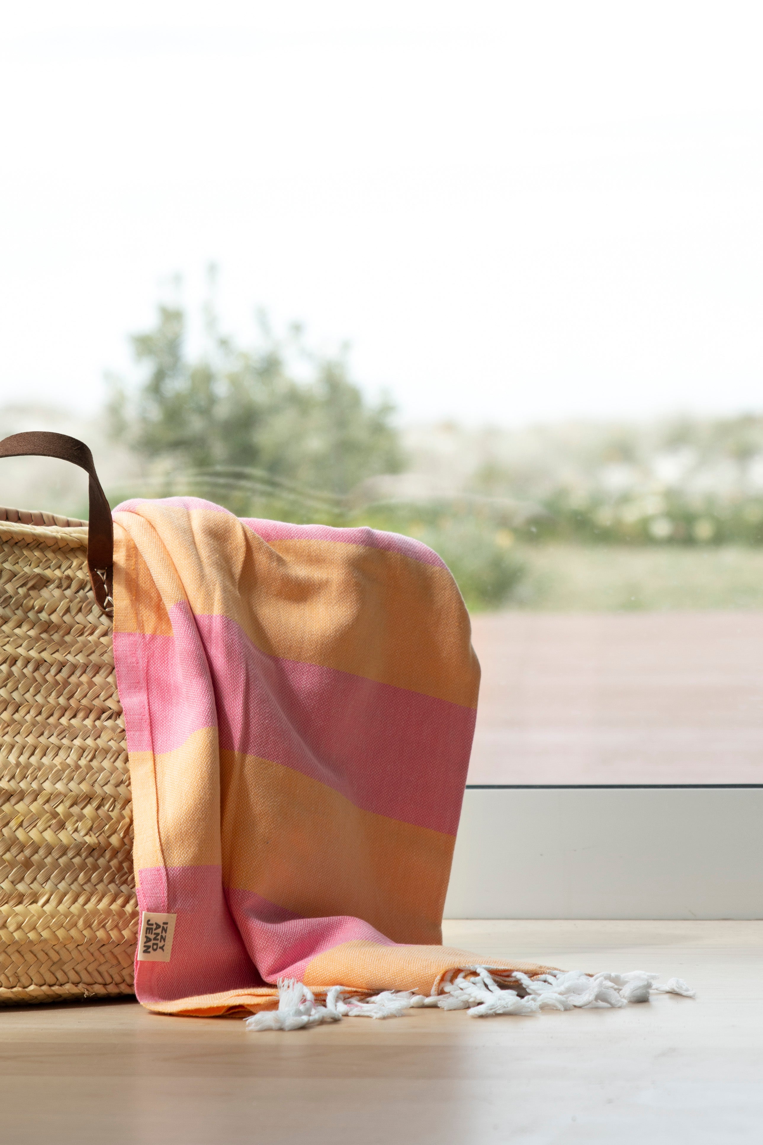 The striped Santorini Turkish towel beach towel in melon and pink hangs over a weave basket near a window overlooking trees