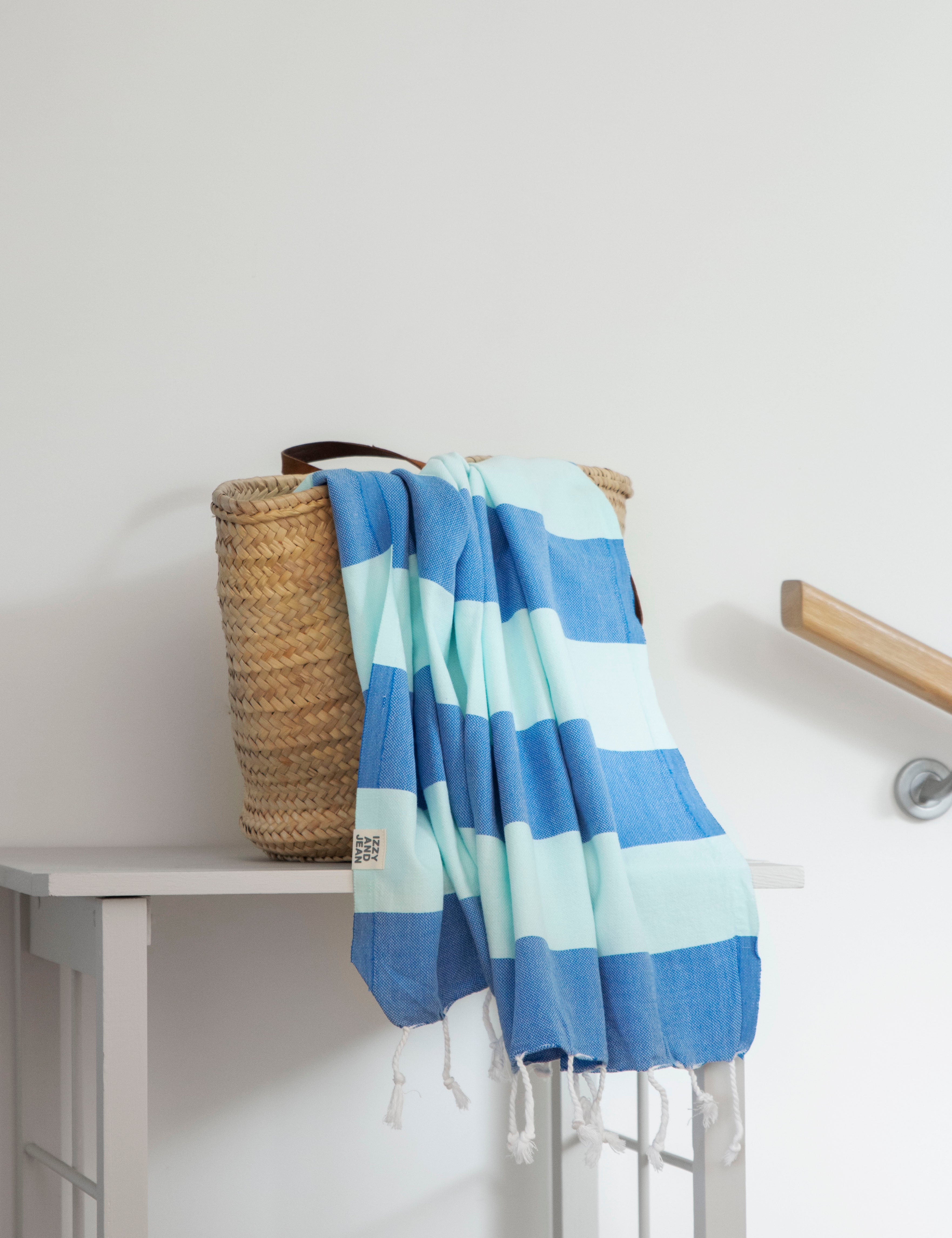 A folded striped towel with bands of blue and mint colours, hangs over a weave basket on a white table