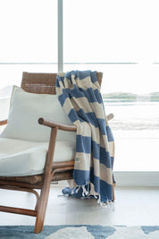 A striped Santorini Turkish towel with navy and sand colours, with white fringes on the edges hangs over the edge of a stylish rattan chair with white pillows