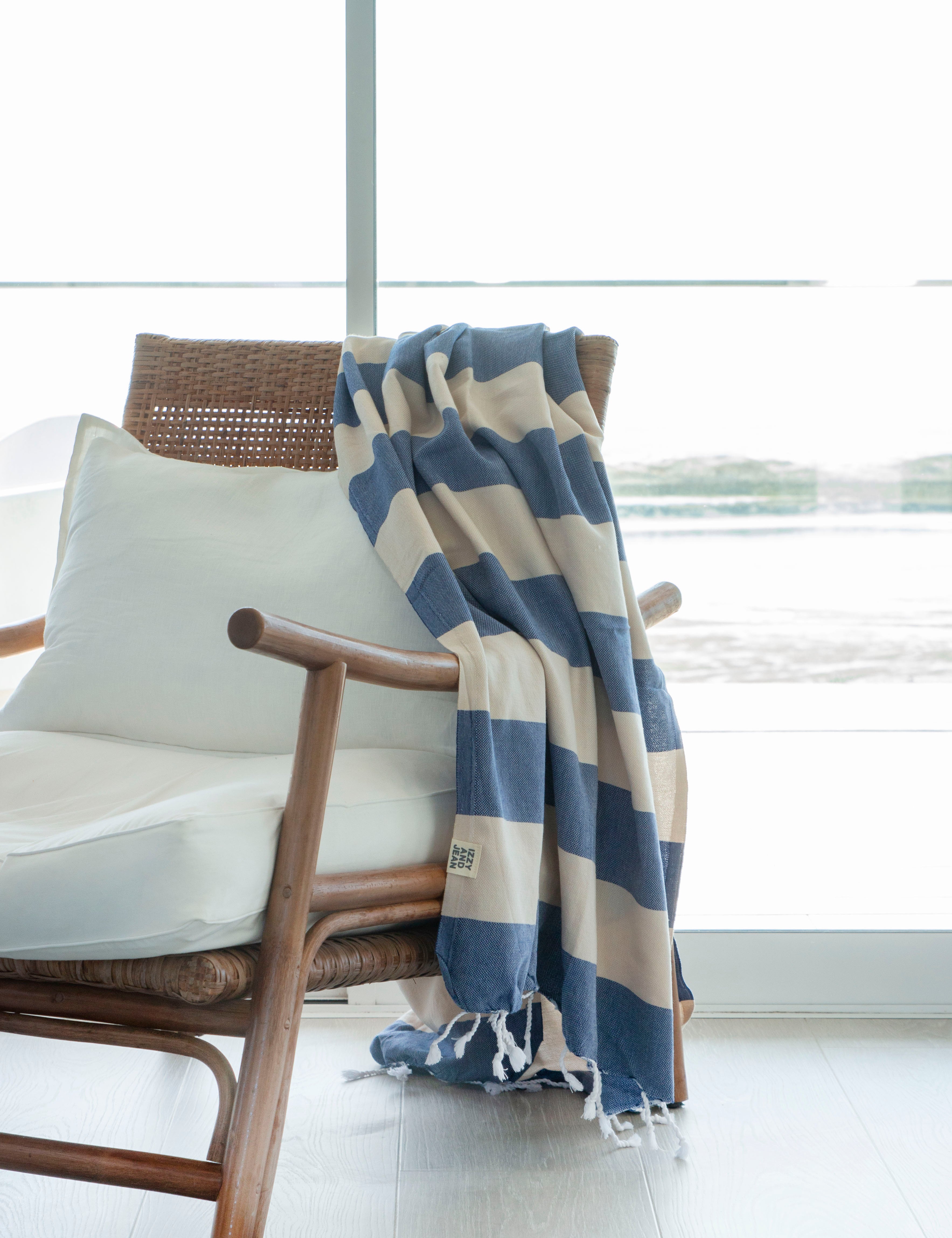 A striped Santorini Turkish towel with navy and sand colours, with white fringes on the edges hangs over the edge of a stylish rattan chair with white pillows