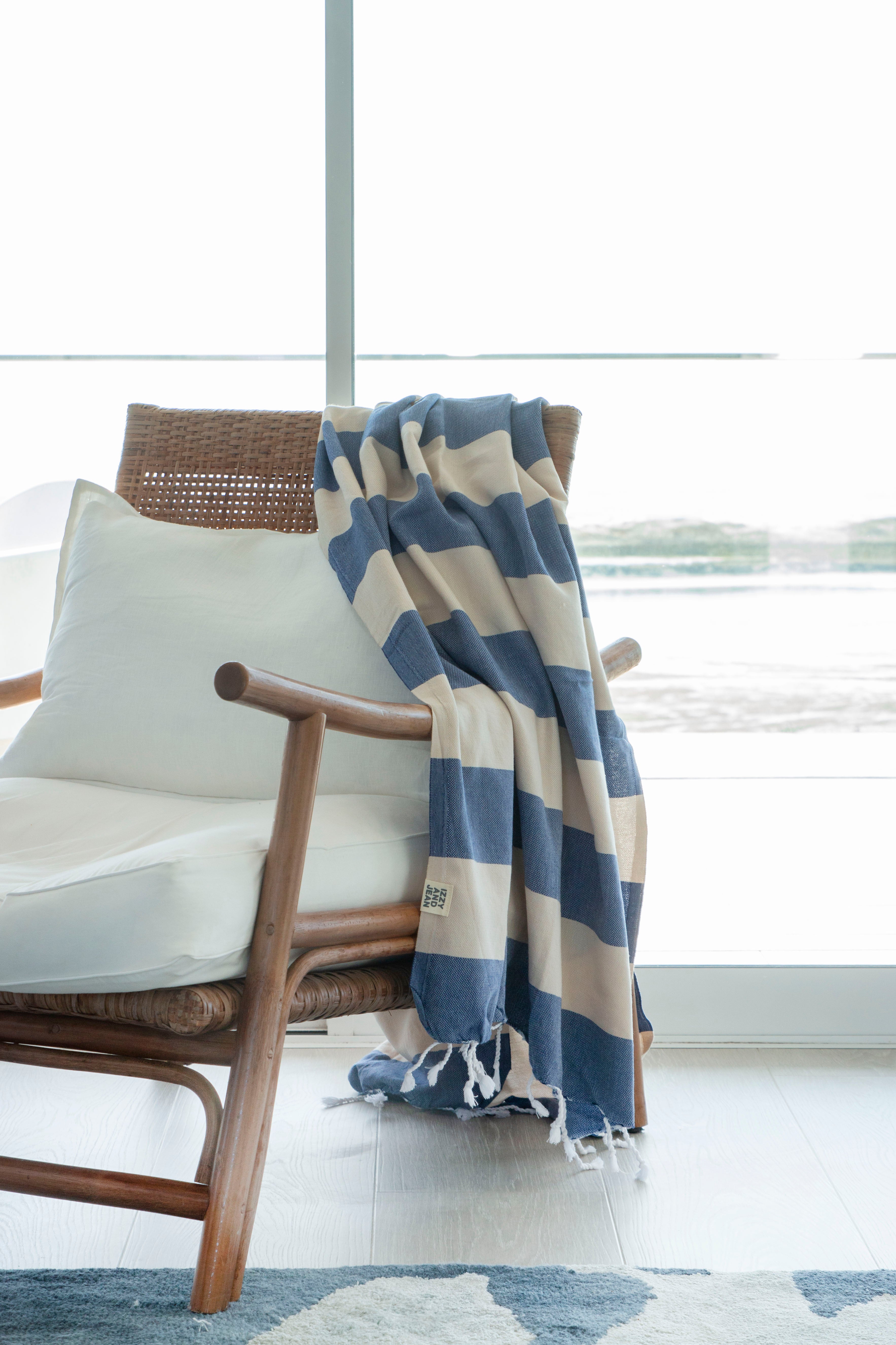 A striped Santorini Turkish towel with navy and sand colours, with white fringes on the edges hangs over the edge of a stylish rattan chair with white pillows