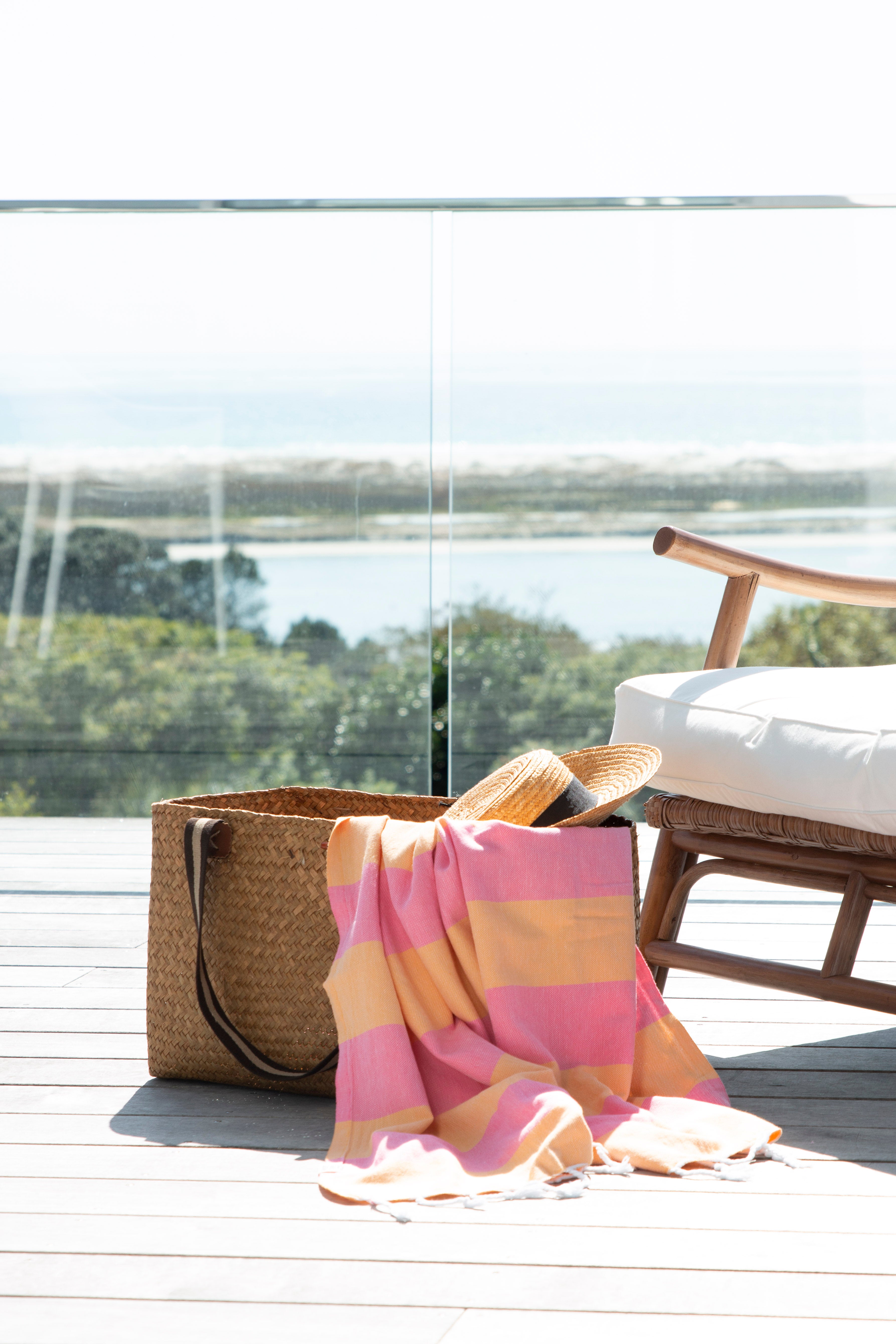 A striped Santorini Turkish towel beach towel in melon and pink, hangs out of a weave basket on a deck overlooking a beach vista next to a sun chair