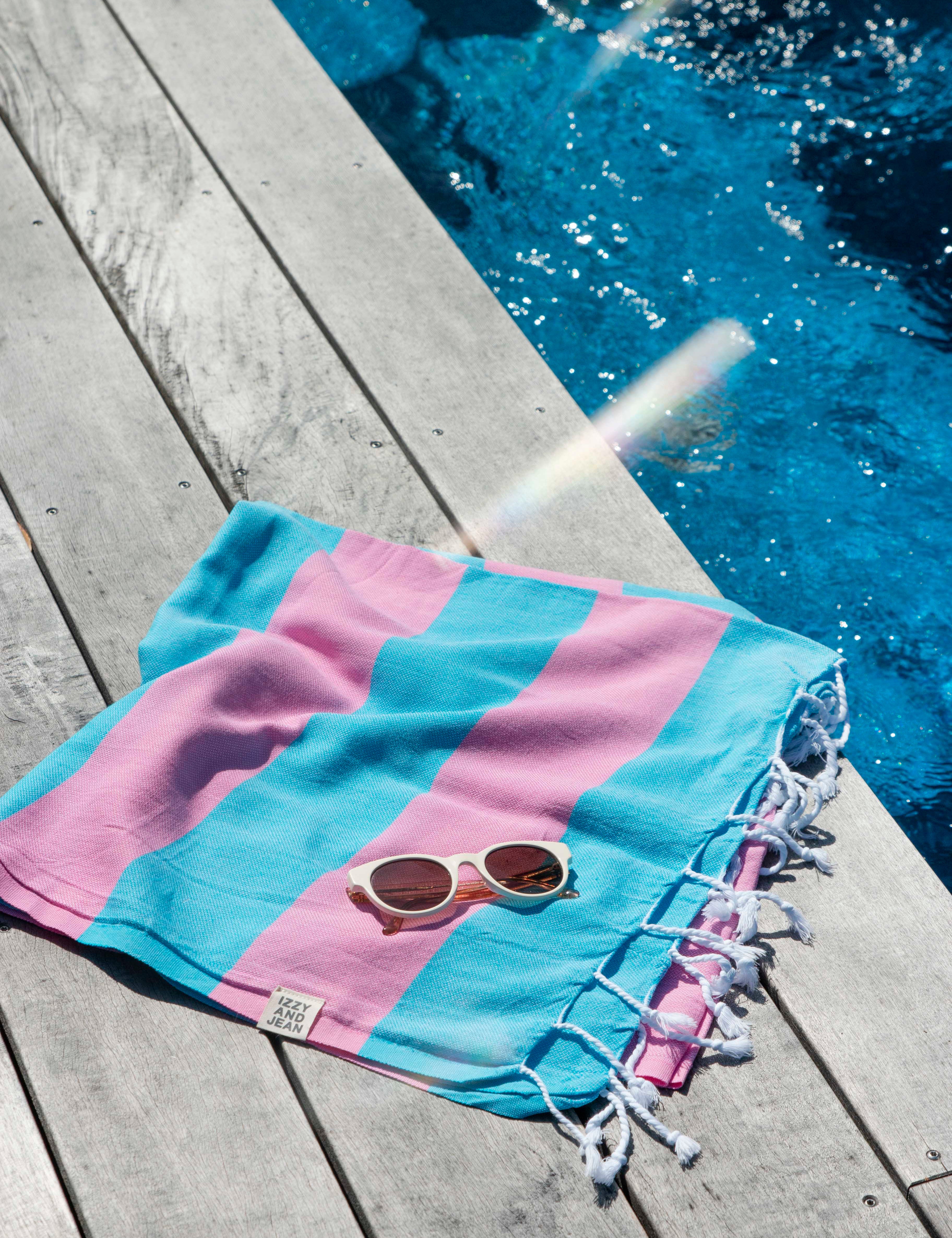 A folded Santorini Turkish towel with marine blue and hot pink stripes with white sunglasses on top is placed beside a pool on a wooden deck 